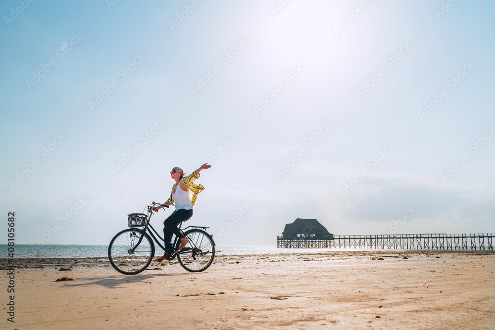 Fototapeta premium Young female dressed light summer clothes joyfully threw up her hand riding old vintage bicycle with front basket on the low tide ocean white sand coast on Kiwengwa beach on Zanzibar island, Tanzania.