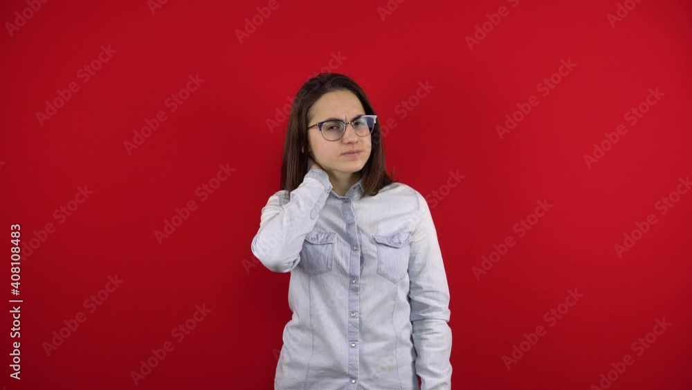 A young woman with glasses has a pain in her neck, she holds her neck with her hand. Shooting on a red background. A young woman with glasses has a pain in her neck, she holds her neck with her hand. Shooting on a red background.