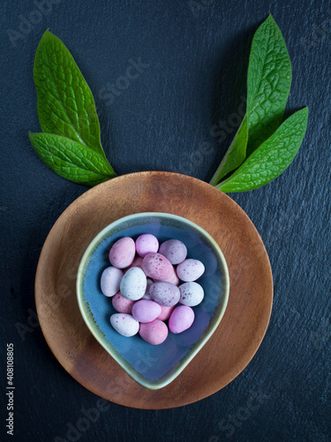 Easter mini chocolate eggs with pink tulips, dark blue slate background. Creative idea, bunny shape.