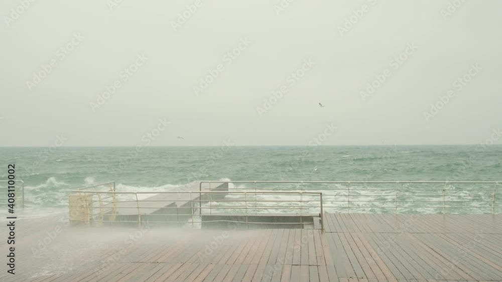 the sea promenade is made of terraced boards, the sea is stormy, waves with foam beat on the pier. Cloudy, seagulls are flying, wide footage the sea promenade is made of terraced boards, the sea is stormy, waves with foam beat on the pier. Cloudy, seagulls are flying, wide footage