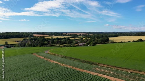 Drone shot approaching group of farmhouses surrounded by trees and fields on a sunny day.