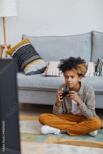Vertical full length portrait of focused African-American boy playing video games at home and holding gamepad while sitting on floor, copy space