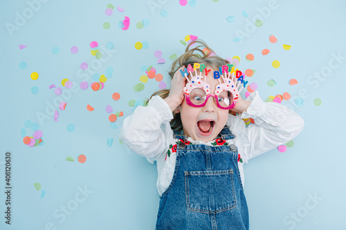 Little girl in happy birthday glasses making funny face, lies on a blue-surfaced floor with colorful confetti pieces. Top view. 