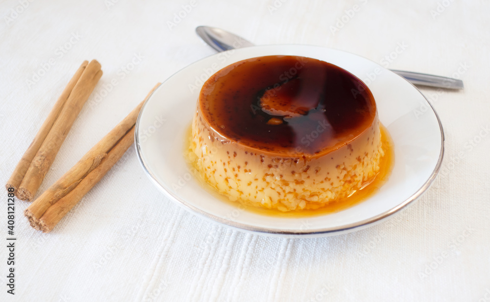 Close-up of a homemade custard pudding on a white plate and some cinnamon sticks, with a spoon ready to eat. Close-up of a homemade custard pudding on a white plate and some cinnamon sticks, with a spoon ready to eat.