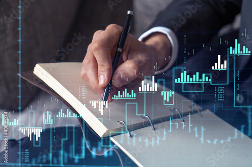 A trader in formal wear writing down some ideas to research stock market to proceed right investment solutions. Wealth management concept. Hologram Forex chart over close up shot.