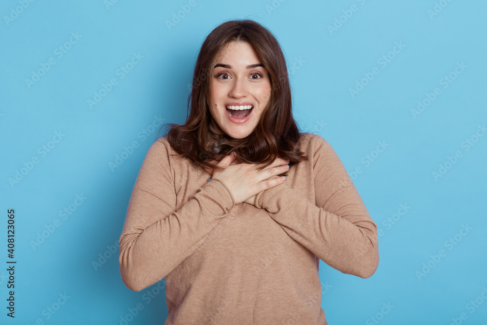 Close up portrait of surprised and impressed wondered brunette girl in beige shirt, gasping amazed, keeps mouth opened, laughing happily, pouting astonished, holds hands on chest, looks excited. Close up portrait of surprised and impressed wondered brunette girl in beige shirt, gasping amazed, keeps mouth opened, laughing happily, pouting astonished, holds hands on chest, looks excited.