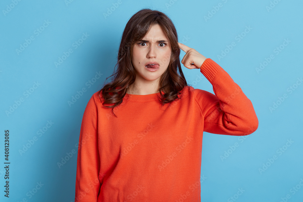 Pretty young caucasian woman isolated on blue background, pointing temple with finger, thinking, focused on task, lady being deeply in thoughts, looking at camera with concentrated look, shows tongue. Pretty young caucasian woman isolated on blue background, pointing temple with finger, thinking, focused on task, lady being deeply in thoughts, looking at camera with concentrated look, shows tongue.