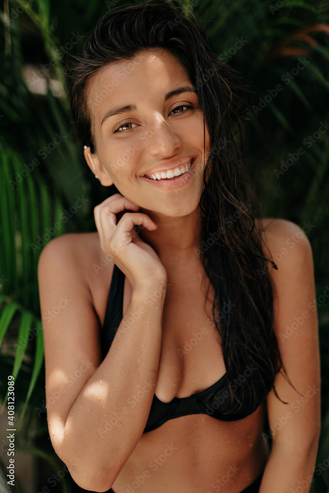 Incredible pretty woman with dark wet hair wearing swimming suit posing with lovable smile on the background of exotic plants Incredible pretty woman with dark wet hair wearing swimming suit posing with lovable smile on the background of exotic plants