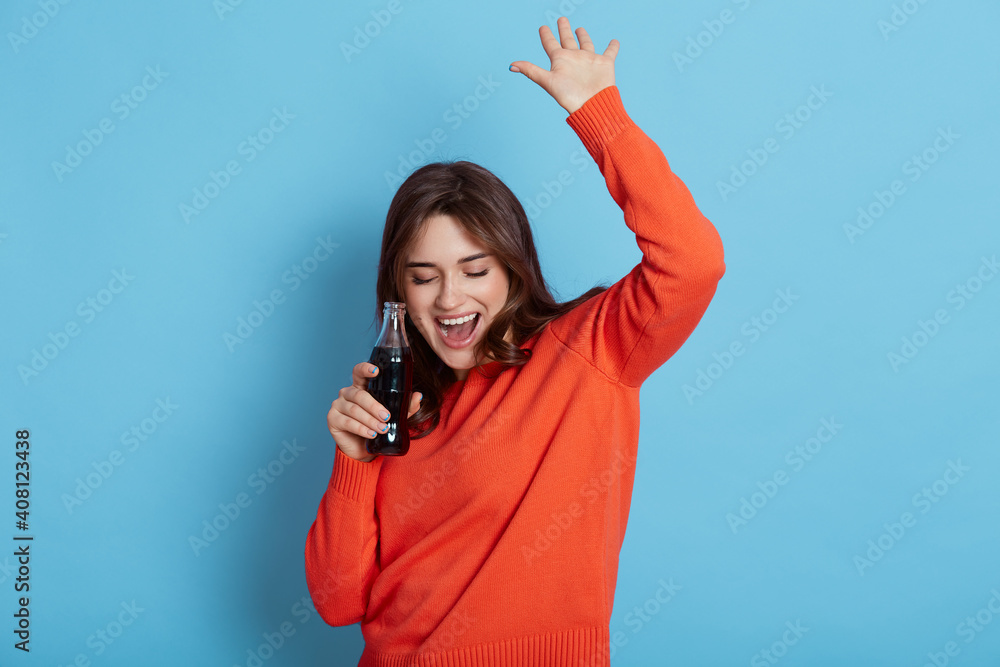 Fototapeta premium Happy crazy brunette girl in casual jumper, holding bottle with soda in hands and dancing happily isolated over blue background, keeping hands raised, yelling something, celebrating good event.,