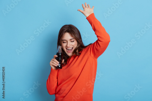 Happy crazy brunette girl in casual jumper, holding bottle with soda in hands and dancing happily isolated over blue background, keeping hands raised, yelling something, celebrating good event.,