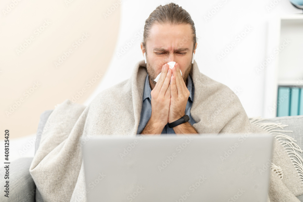 The guy has a cold and flu, he sneezes into paper tissue sitting at home covered with blanket. A young man has viral illness worried about his sick, sitting om the couch with a laptop The guy has a cold and flu, he sneezes into paper tissue sitting at home covered with blanket. A young man has viral illness worried about his sick, sitting om the couch with a laptop