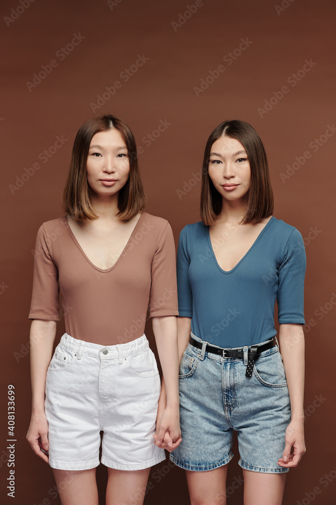 Young twin sisters in casual pullovers and shorts standing close to one another Young twin sisters in casual pullovers and shorts standing close to one another