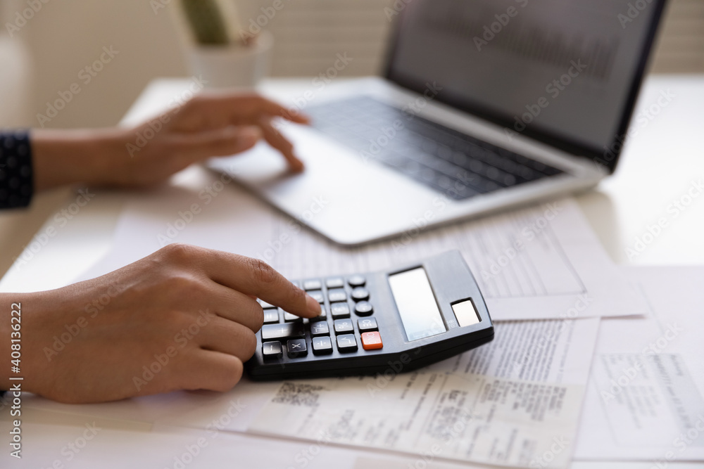 Close up of young female electronic bank client using calculator in paperwork with accounts bills before providing payment online by laptop. Woman hands at work table with papers accounting sum to pay Close up of young female electronic bank client using calculator in paperwork with accounts bills before providing payment online by laptop. Woman hands at work table with papers accounting sum to pay