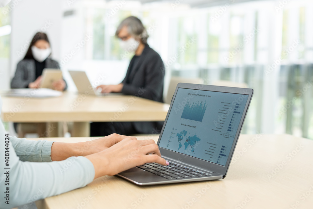 Hands of young businesswoman using laptop by desk against two busy colleagues Hands of young businesswoman using laptop by desk against two busy colleagues