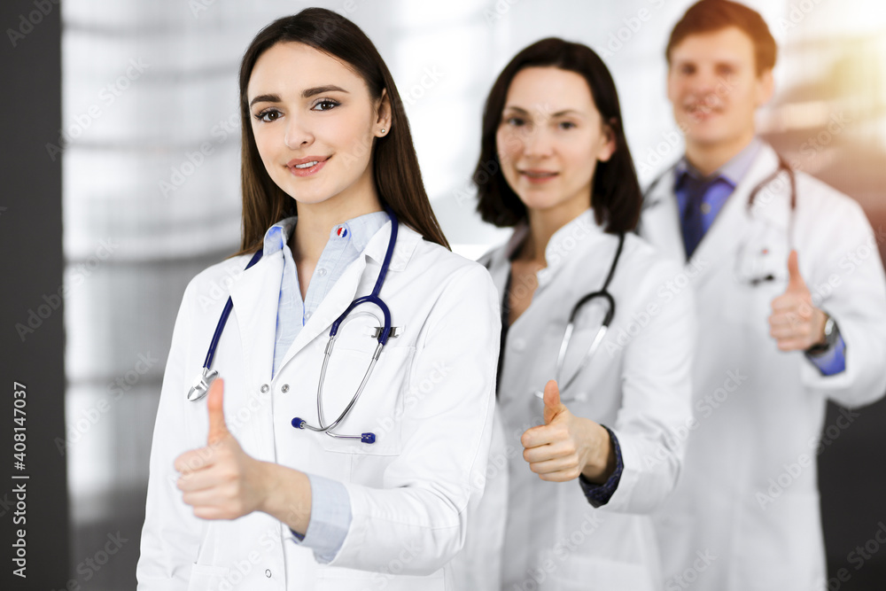 Group of young cheerful doctors standing as a team with thumbs up in sunny hospital. Medical help, insurance in health care, best disease treatment and medicine concept Group of young cheerful doctors standing as a team with thumbs up in sunny hospital. Medical help, insurance in health care, best disease treatment and medicine concept