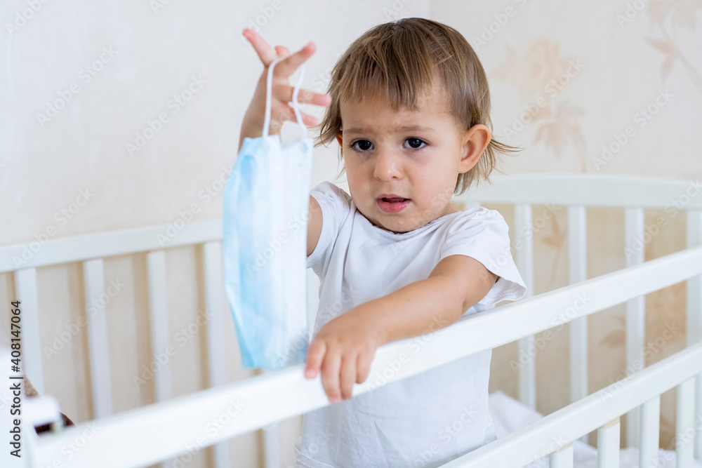 coronavirus covid-19 quarantine concept, precautions. little cute toddler stands in the crib at home and holds out a medical mask coronavirus covid-19 quarantine concept, precautions. little cute toddler stands in the crib at home and holds out a medical mask