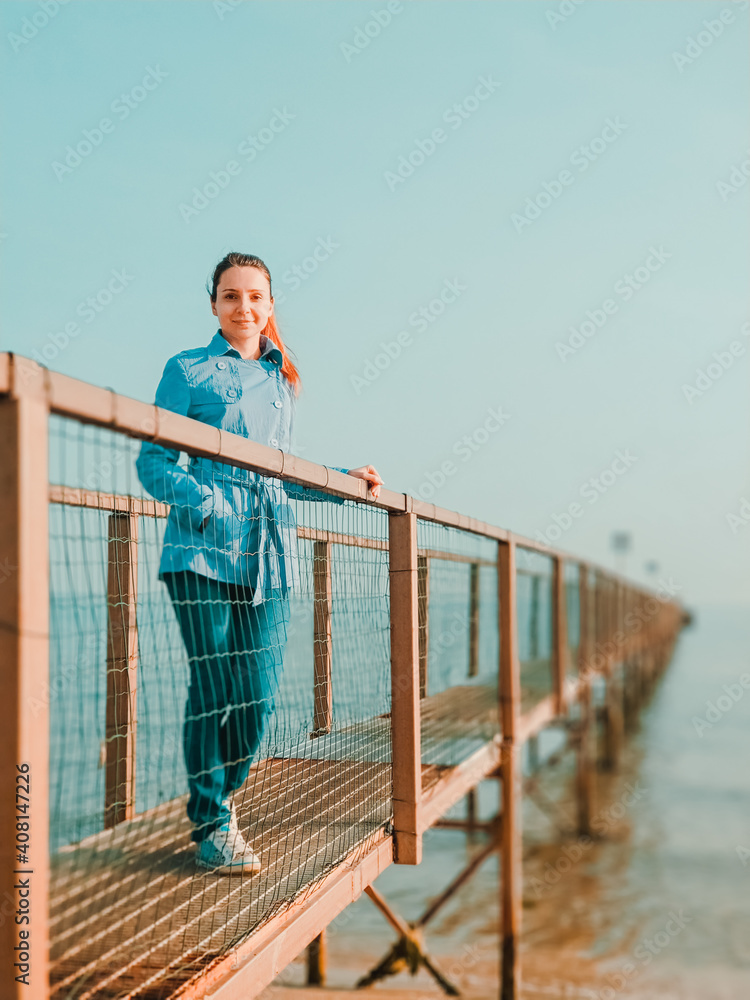 Girl in casual jacket and jeans walking on a pier on the beach. Young woman enjoying beauty of nature while walking on the sea bridge and looking at camera Girl in casual jacket and jeans walking on a pier on the beach. Young woman enjoying beauty of nature while walking on the sea bridge and looking at camera