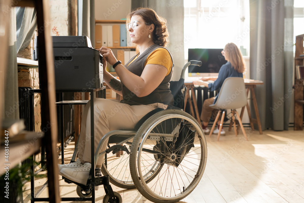 Young serious disable businesswoman in wheelchair sitting by xerox machine Young serious disable businesswoman in wheelchair sitting by xerox machine