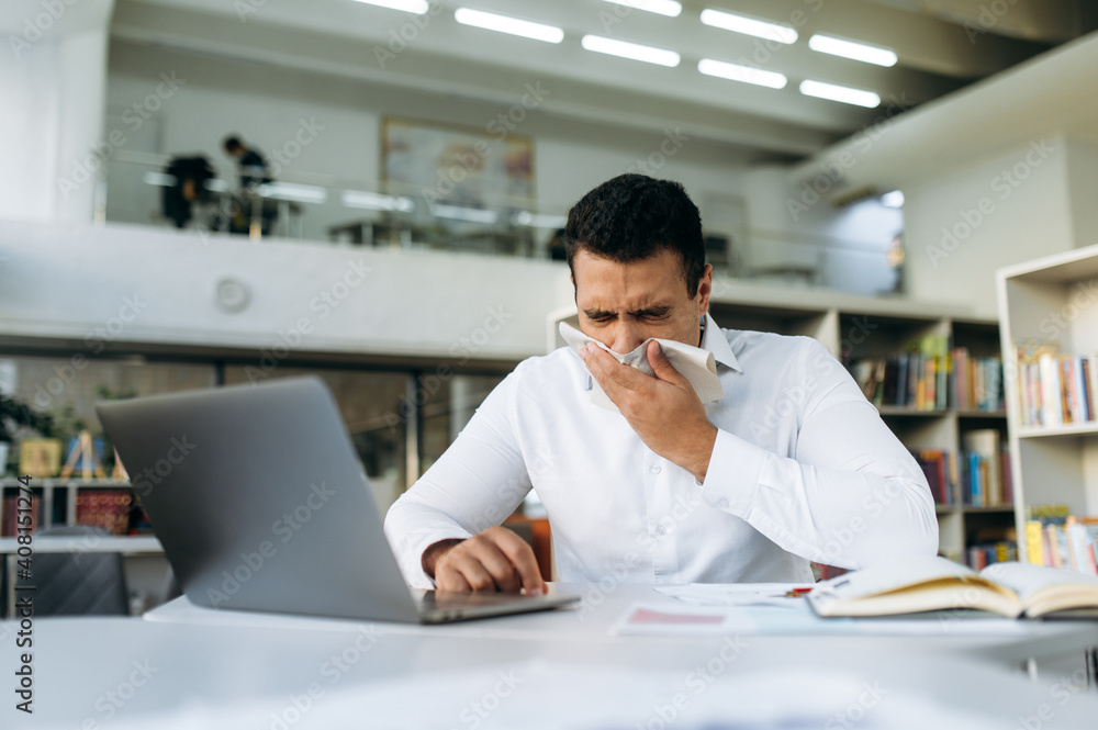Hispanic business man sneezing, using a paper tissue, feeling unwell. Unhealthy manager struggling with running nose, catch a flu, remotely working or studying, while illness Hispanic business man sneezing, using a paper tissue, feeling unwell. Unhealthy manager struggling with running nose, catch a flu, remotely working or studying, while illness
