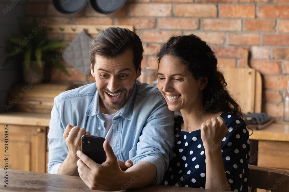 Fototapeta premium Excited millennial Caucasian man and woman feel euphoric win online lottery on smartphone. Happy overjoyed young couple look at cellphone screen triumph with good news or deal message.