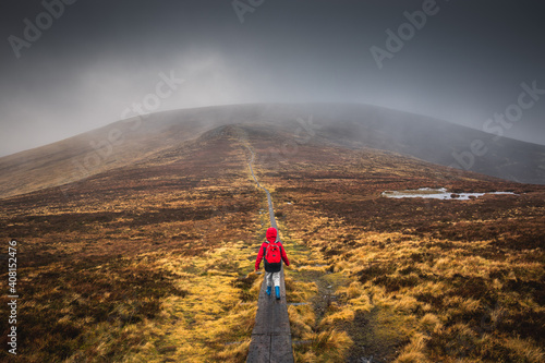 Boy in a red jacket, hiking on wooden path leading through the wicklow mountains, Djouce pek Ireland. Wooden path in foggy mountain landscape, in Autumn. 2019 Irland