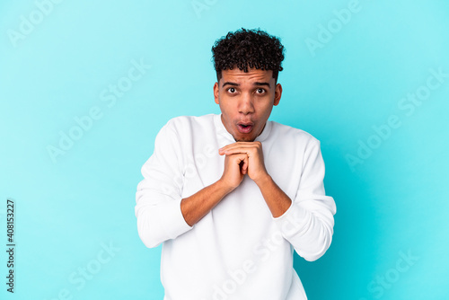 Young african american curly man isolated on blue praying for luck, amazed and opening mouth looking to front.