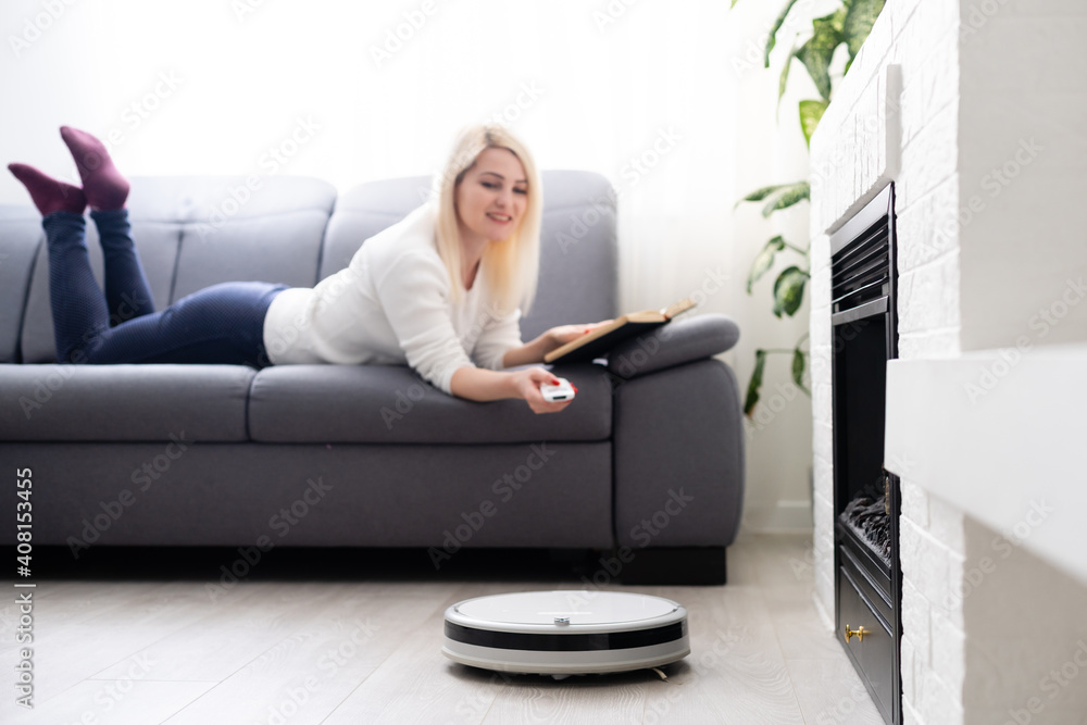 Robotic vacuum cleaner cleaning the room while woman relaxing on sofa. Woman controlling vacuum with remote control Robotic vacuum cleaner cleaning the room while woman relaxing on sofa. Woman controlling vacuum with remote control