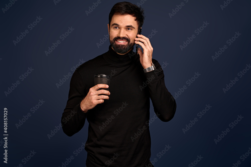 Smiling young man in trendy sweater posing isolated on Blue wall background studio portrait. Guy making phone call standing, holding paper coffee cup or tea Smiling young man in trendy sweater posing isolated on Blue wall background studio portrait. Guy making phone call standing, holding paper coffee cup or tea