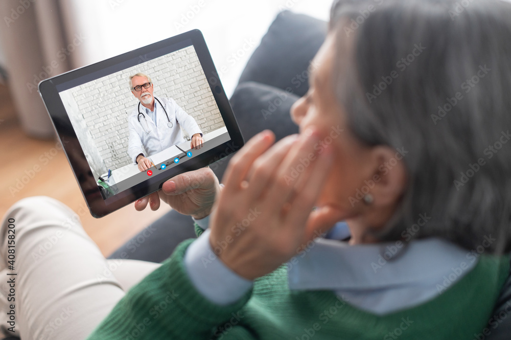 Close-up of a senior mature lady wearing a green sweater holding a tablet and talking with a therapist doctor, getting consultation about her health condition, cyber medicine or self-isolation concept Close-up of a senior mature lady wearing a green sweater holding a tablet and talking with a therapist doctor, getting consultation about her health condition, cyber medicine or self-isolation concept