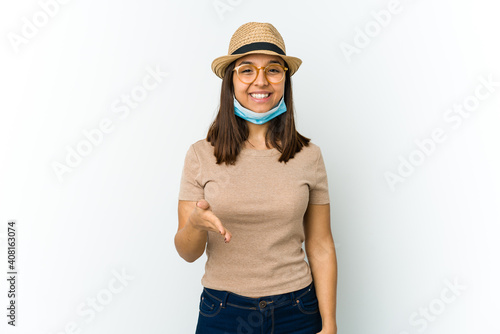 Young latin woman wearing hat and mask to protect from covid isolated on white background stretching hand at camera in greeting gesture.
