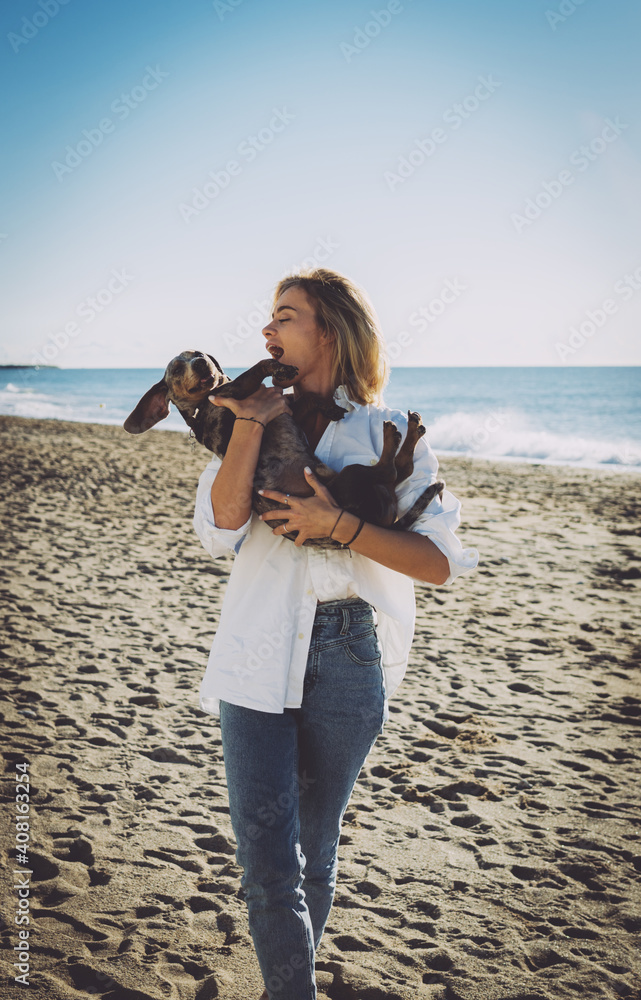 Cheerful blonde female hugging her lovely pet dog while standing at the beach on a sea shore background. Attractive Caucasian female in white shirt and blue jeans enjoying the day off with her doggy. Cheerful blonde female hugging her lovely pet dog while standing at the beach on a sea shore background. Attractive Caucasian female in white shirt and blue jeans enjoying the day off with her doggy.