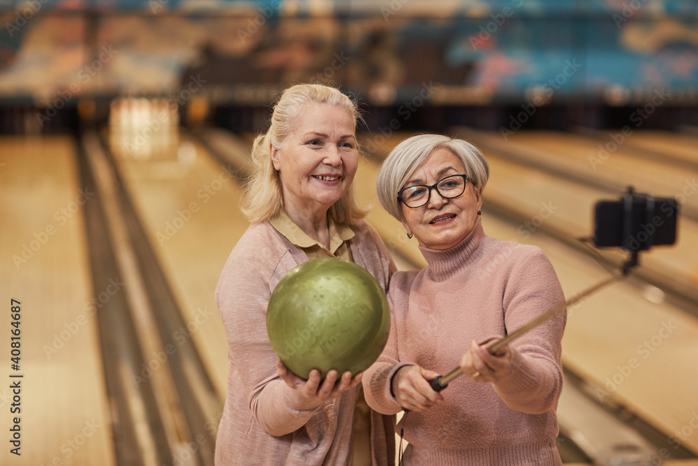Fototapeta premium Waist up portrait of two senior women taking selfie photos while playing bowling and enjoying active entertainment at bowling alley, copy space