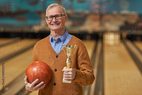 Waist up portrait of smiling senior man holding trophy and bowling ball while posing at bowling alley after winning match, copy space