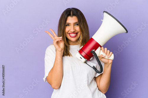 Young indian woman holding a megaphone isolated joyful and carefree showing a peace symbol with fingers.