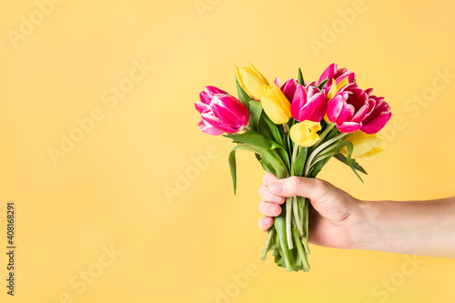 Male hand holding beautiful fresh pink and yellow tulips bouquet on light yellow background. Greeting card mockup. Birthday, Valentines day, mother's and woman's day. Selective focus. Copy space.