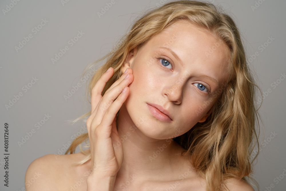 Closeup of pretty young woman face with blue eyes, curly natural blonde  hair, has no makeup, touching her soft skin, standing shirtless with bare  shoulders, looking at camera. Studio grey background Photos