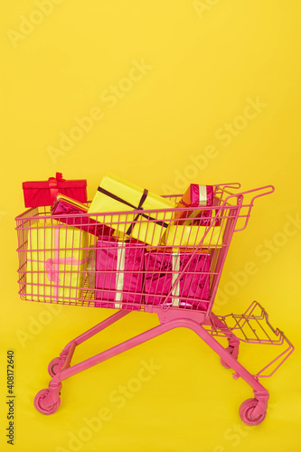 Gift buying. Shopping cart full of boxes. Pink trolley with presents isolated on yellow background. Christmas, Xmas, birthday gift. Concept of celebration, retail and sale. Online Shopping. copy space