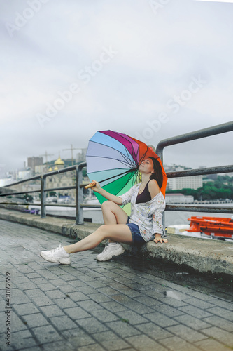 young beautiful woman in floral print shirt, denim shorts and white large sneakers sits on wet paving stones in rainy weather under rainbow umbrella in an urban atmosphere, vertical content © Aleksandra