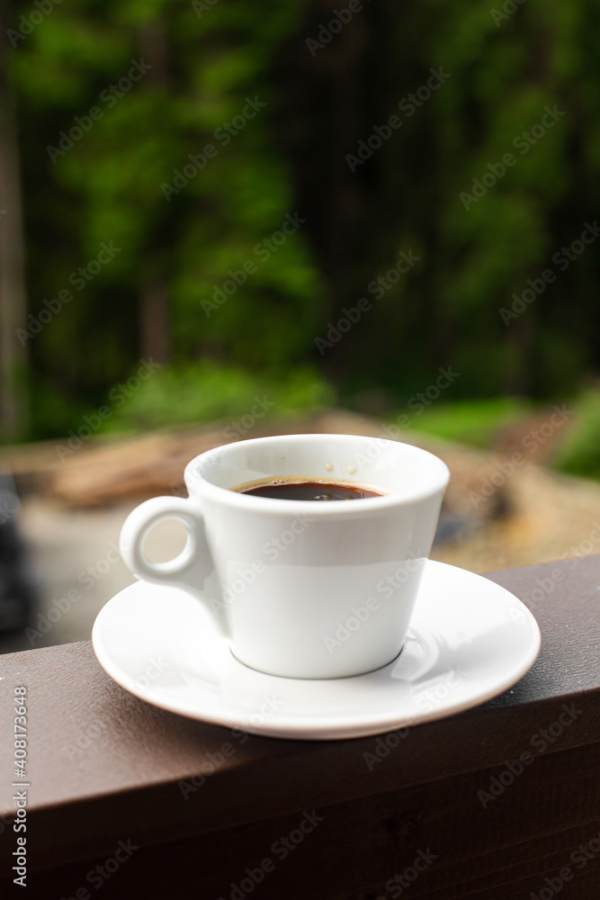 The white mug of dark hot morning coffee on wooden fence with forest background The white mug of dark hot morning coffee on wooden fence with forest background