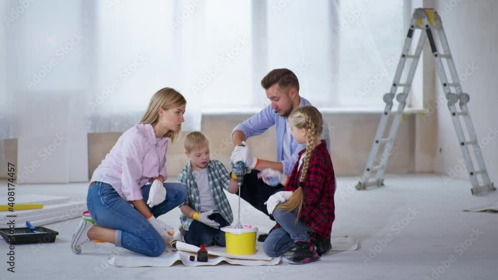 finishing work, cheerful woman with down syndrome girl and boy watch young man mix pigment with paint in bucket using construction drill indoors to paint walls finishing work, cheerful woman with down syndrome girl and boy watch young man mix pigment with paint in bucket using construction drill indoors to paint walls