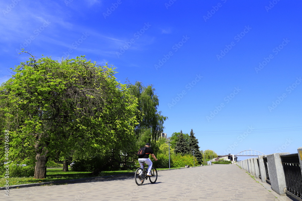 A man rides a bicycle along the embankment by the river, summer street, boulevard with large trees against the blue sky. Spring street, Dnipro, Dnepropetrovsk, Ukraine