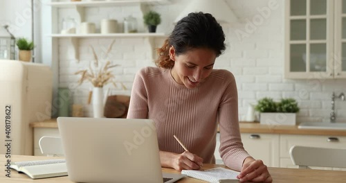 Happy young woman web surfing educational information in internet using computer app, preparing for exams or studying on online courses sitting at table in modern kitchen, writing notes in copybook. photo