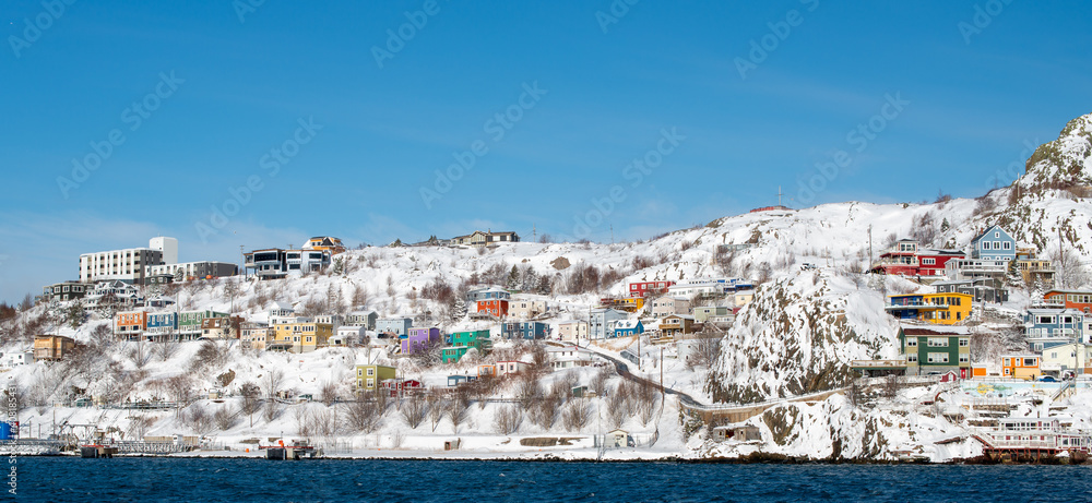 The historic Battery neighbourhood in St. John's, Newfoundland after a blanket of snow.  The bright and colourful houses are built on the hillside. There's a blue sky and deep blue ocean near the hill