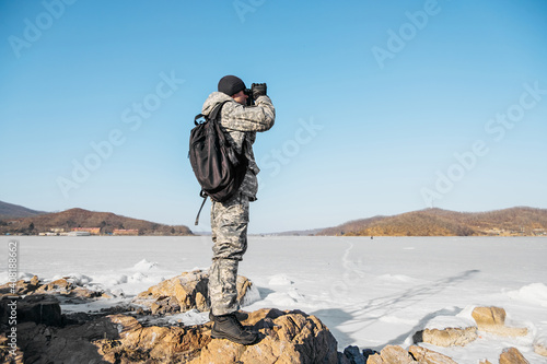 man in military camouflage uniform, black hat and backpack, looks through binoculars at frozen sea around. winter local travel, selective focus photo