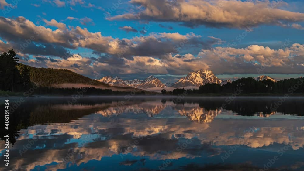 Wide view of beautiful pink and golden sunrise at reflective mirror lake with snow capped mountains and thick clouds flowing by in Grand Teton National Park, Wyoming United States Wide view of beautiful pink and golden sunrise at reflective mirror lake with snow capped mountains and thick clouds flowing by in Grand Teton National Park, Wyoming United States