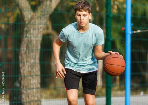 Cute young boy plays basketball on street playground in summer. Teenager in green t shirt with orange basketball ball outside. Hobby, active lifestyle, sport activity for kids. © Natali