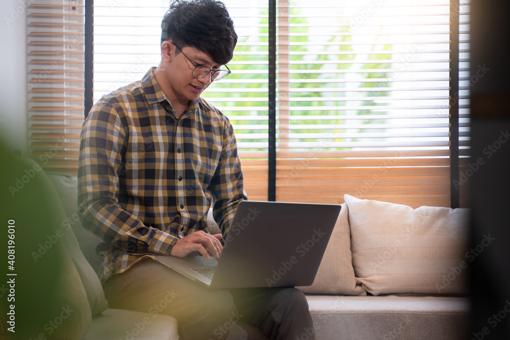 young attractive asian man relaxing comfortably working on laptop in bright living room at his home. young attractive asian man relaxing comfortably working on laptop in bright living room at his home.