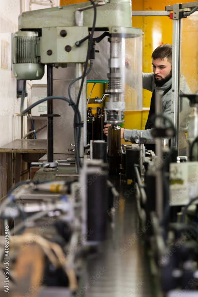 Skilled guy working on conveyor for bottling olive oil in glass bottles in small family factory