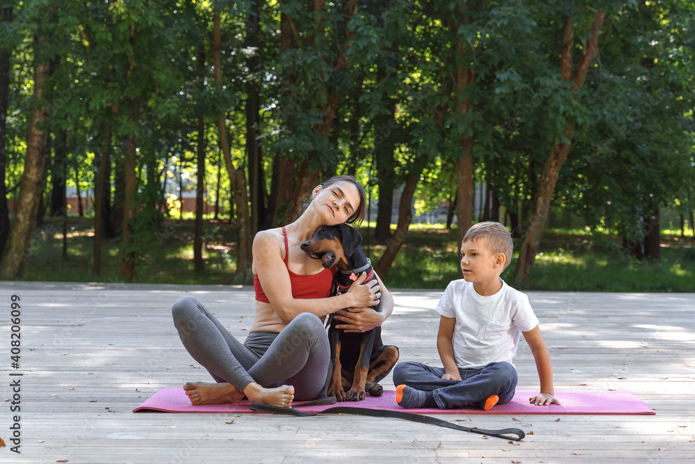 happy family is engaged in gymnastics in a city park with a dog. mom, son and doberman spend time together. family relaxing and doing fitness at nature
