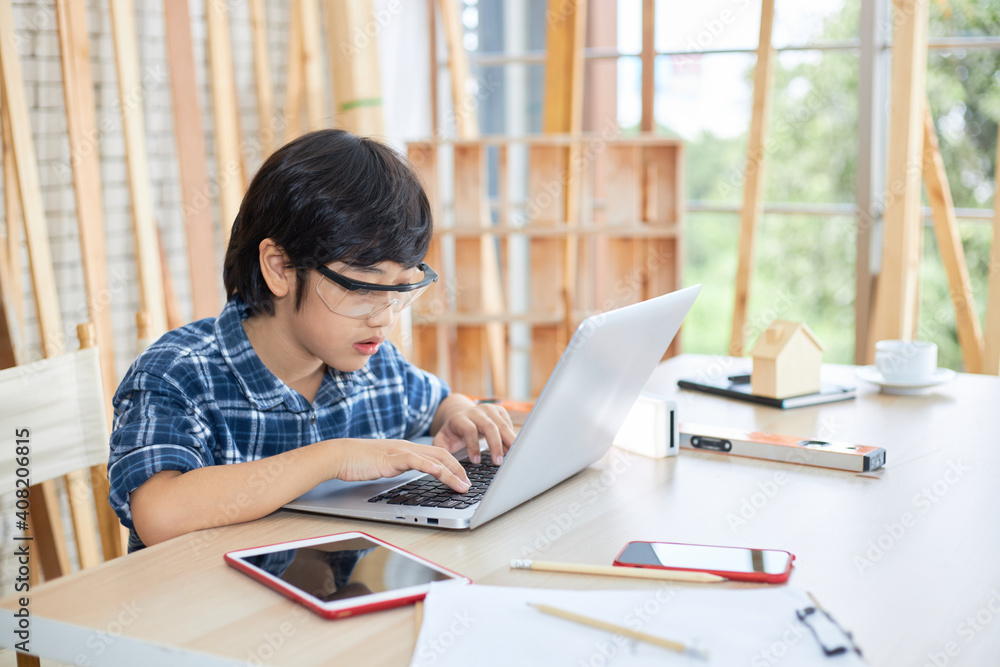Asian boy is using a laptop in a room full of hand tools and piles of wood.